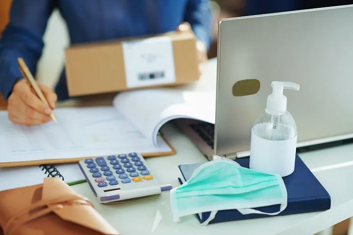 Hand sanitizer, surgical masks, and calculators are all present as a worker checks a shipping manifest