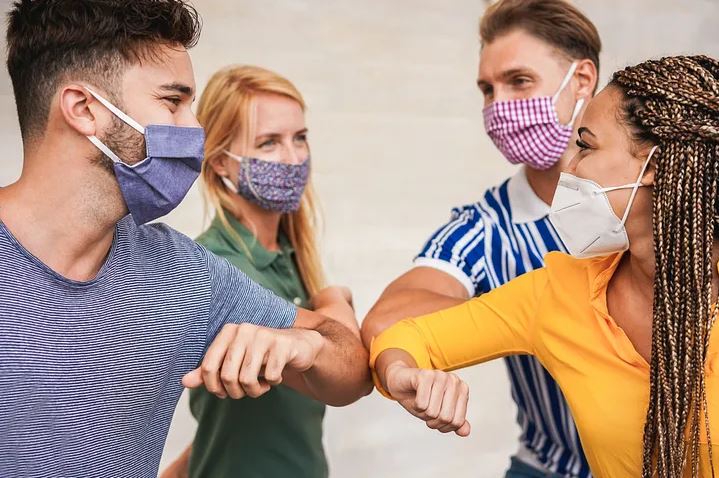 A group of four people bump elbows to acknowledge each other while mitigating germ spread. All group members wear a mask to protect themselves from Covid-19