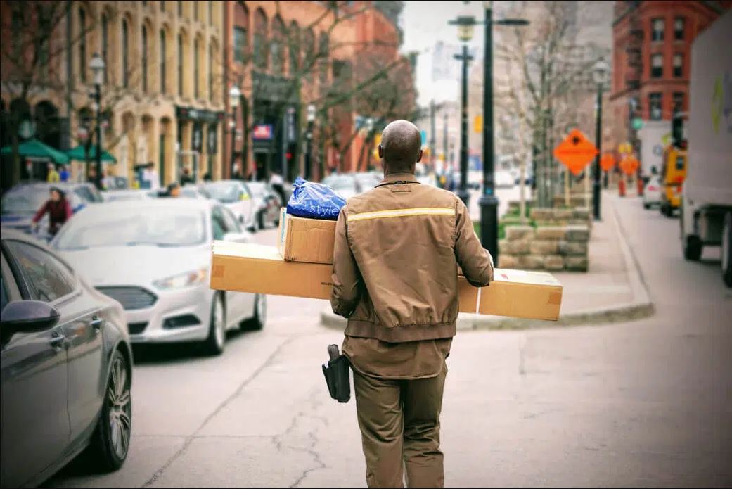 A UPS worker carries several boxes through a busy street to their destination